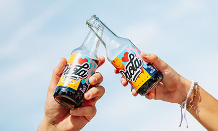 Deux personnes trinquant avec des bouteilles de soda local Paola, sous un ciel bleu